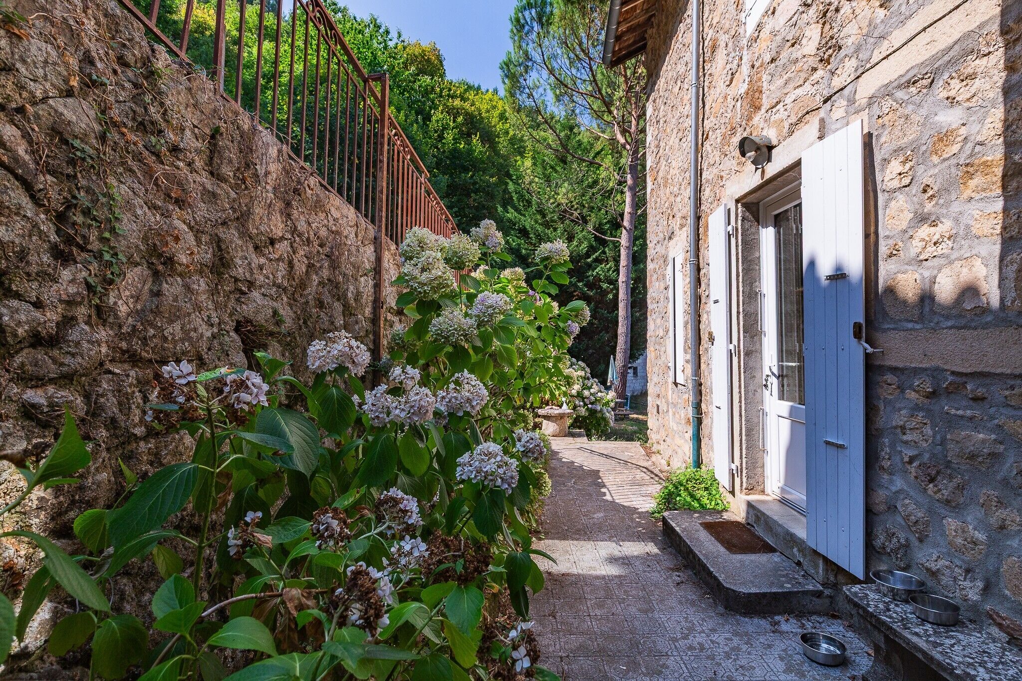 A narrow stone pathway leading to a rustic stone building with a white door, surrounded by lush greenery and flowering plants.