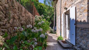 Un étroit sentier de pierres mène à un bâtiment rustique en pierre, doté d’une porte blanche, et est entouré d’une végétation luxuriante et de plantes en fleurs.