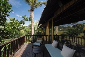Una terraza de madera con mobiliario exterior, rodeada de abundante vegetación y un cielo despejado.