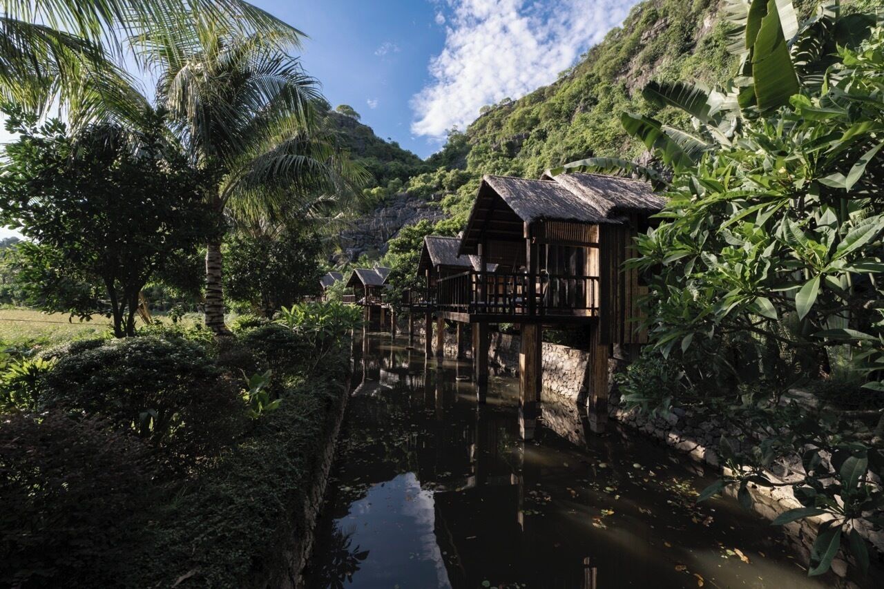 Une structure en bois surmontée d’un toit de chaume, entourée d’une végétation luxuriante et bordée d’une étendue d’eau paisible.