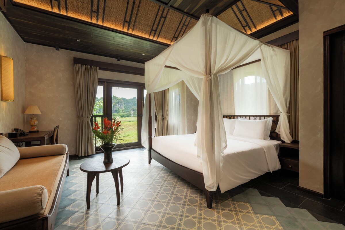 A hotel room with a bed, a sofa, a small table with a vase, and a view of the outdoors.