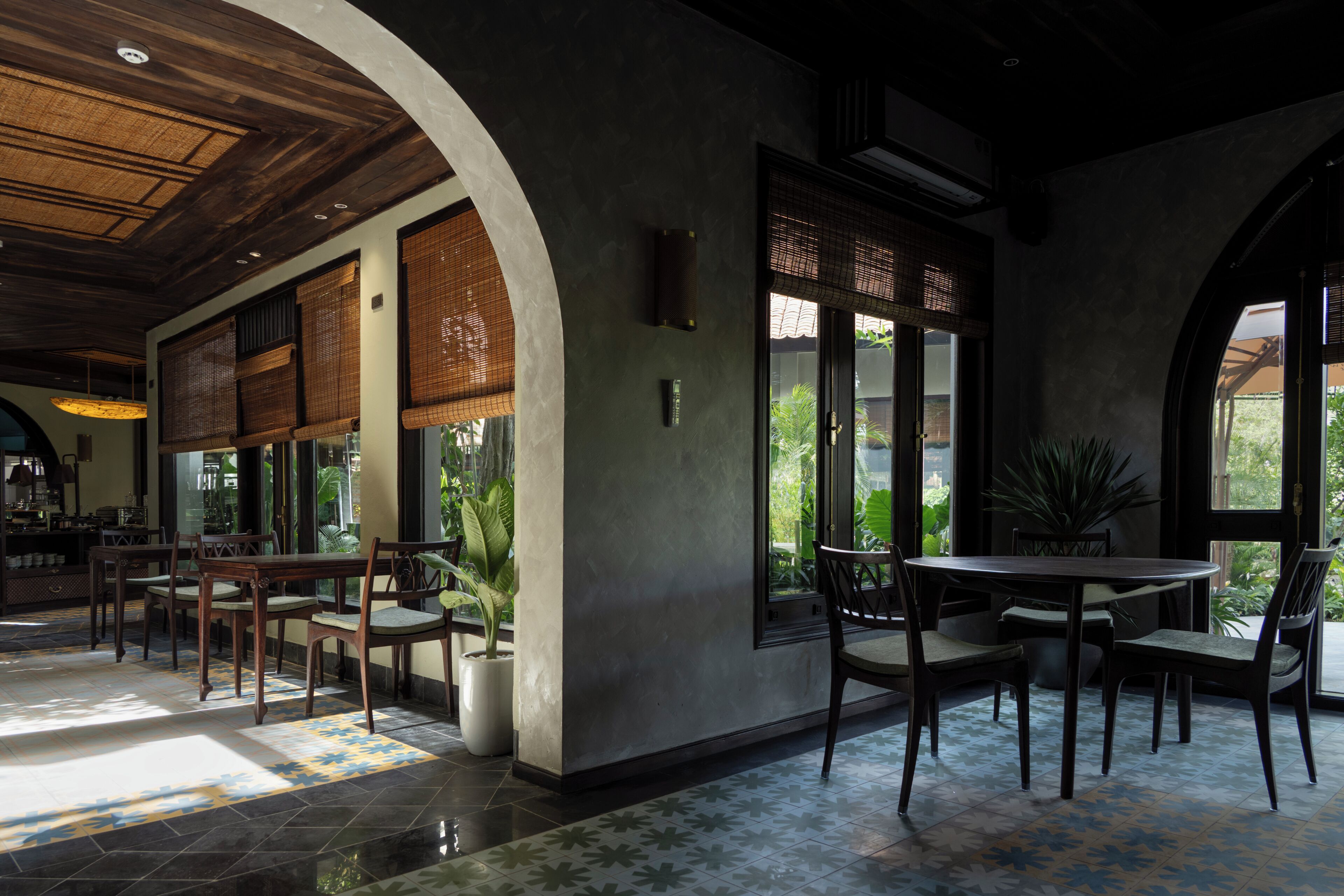 Un comedor interior con mesas y sillas de madera, ventanas amplias y plantas en macetas.