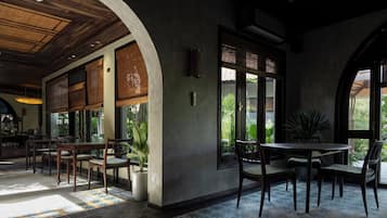 Un comedor interior con mesas y sillas de madera, ventanas amplias y plantas en macetas.