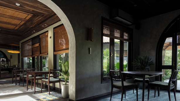 Un comedor interior con mesas y sillas de madera, ventanas amplias y plantas en macetas.
