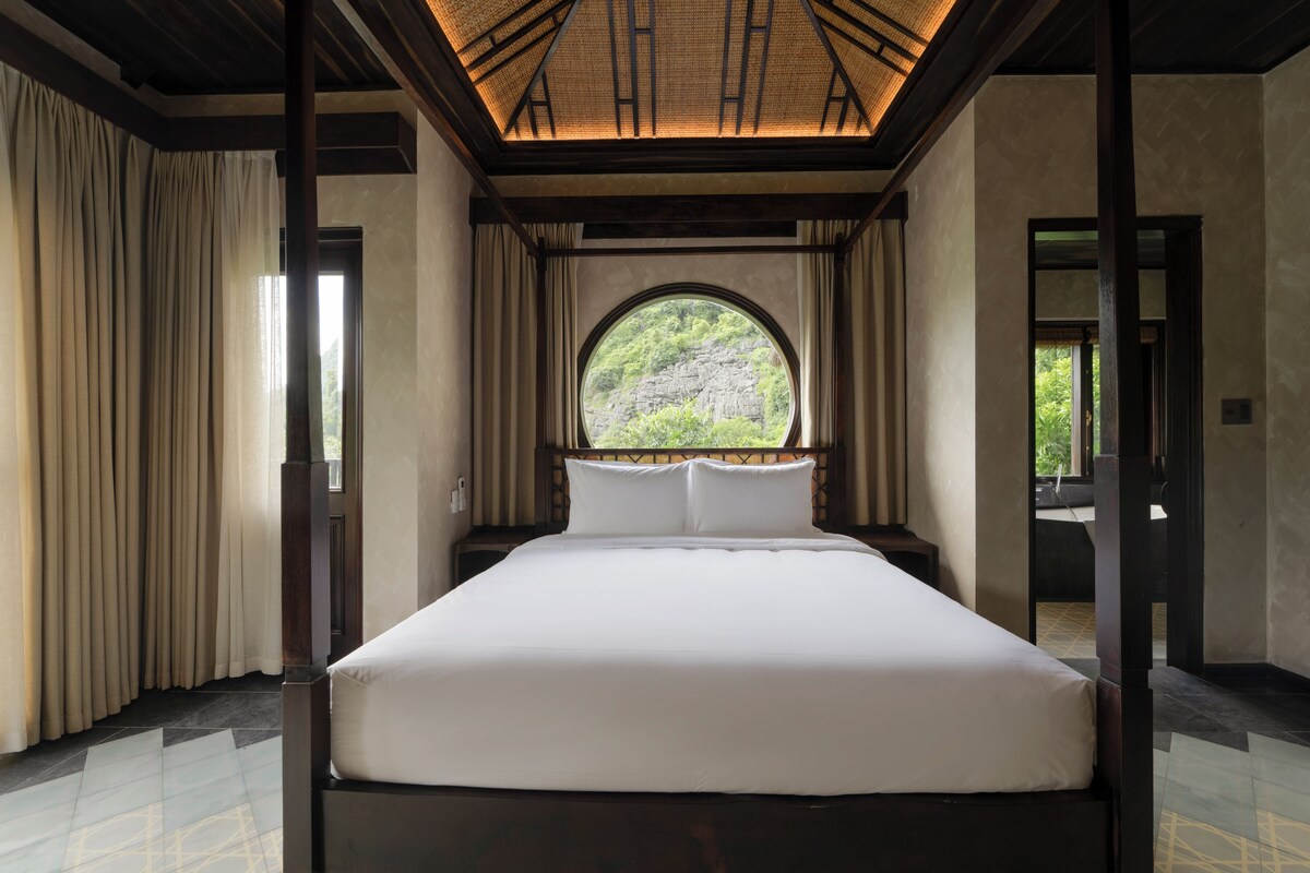 A large bed in a room with a view through a window, curtains, and a wooden ceiling.
