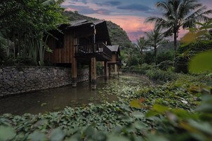 A wooden house with a balcony, surrounded by lush greenery and a body of water.