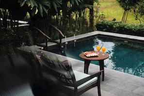 Zona junto a la piscina con una mesa lista para dos personas, una silla y una pequeña mesa con comida y bebidas.