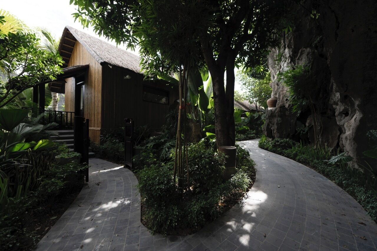 A pathway leading to a wooden building with a thatched roof, surrounded by lush greenery and a rocky cliff.