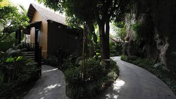 A pathway leading to a wooden building with a thatched roof, surrounded by lush greenery and a rocky cliff.