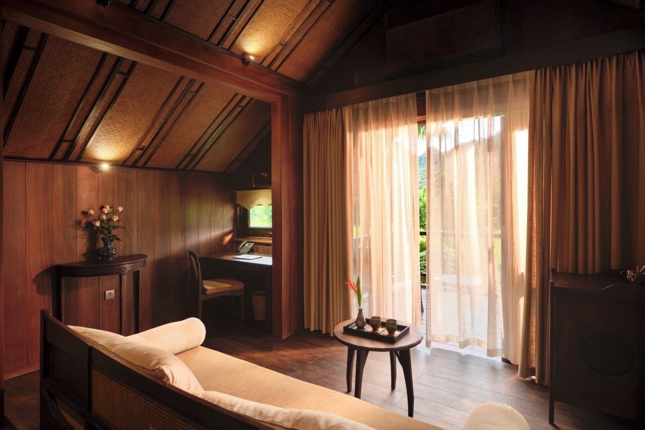 Ein Zimmer mit einem Bett, einem kleinen Tisch mit einer Vase, einem Schreibtisch mit Stuhl und einem großen Fenster mit Vorhängen.