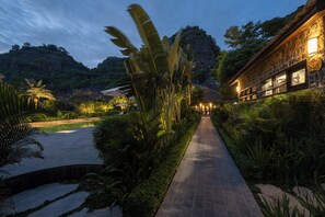 Un camino conduce a un edificio de fachada de piedra, rodeado de exuberante vegetación y una piscina.