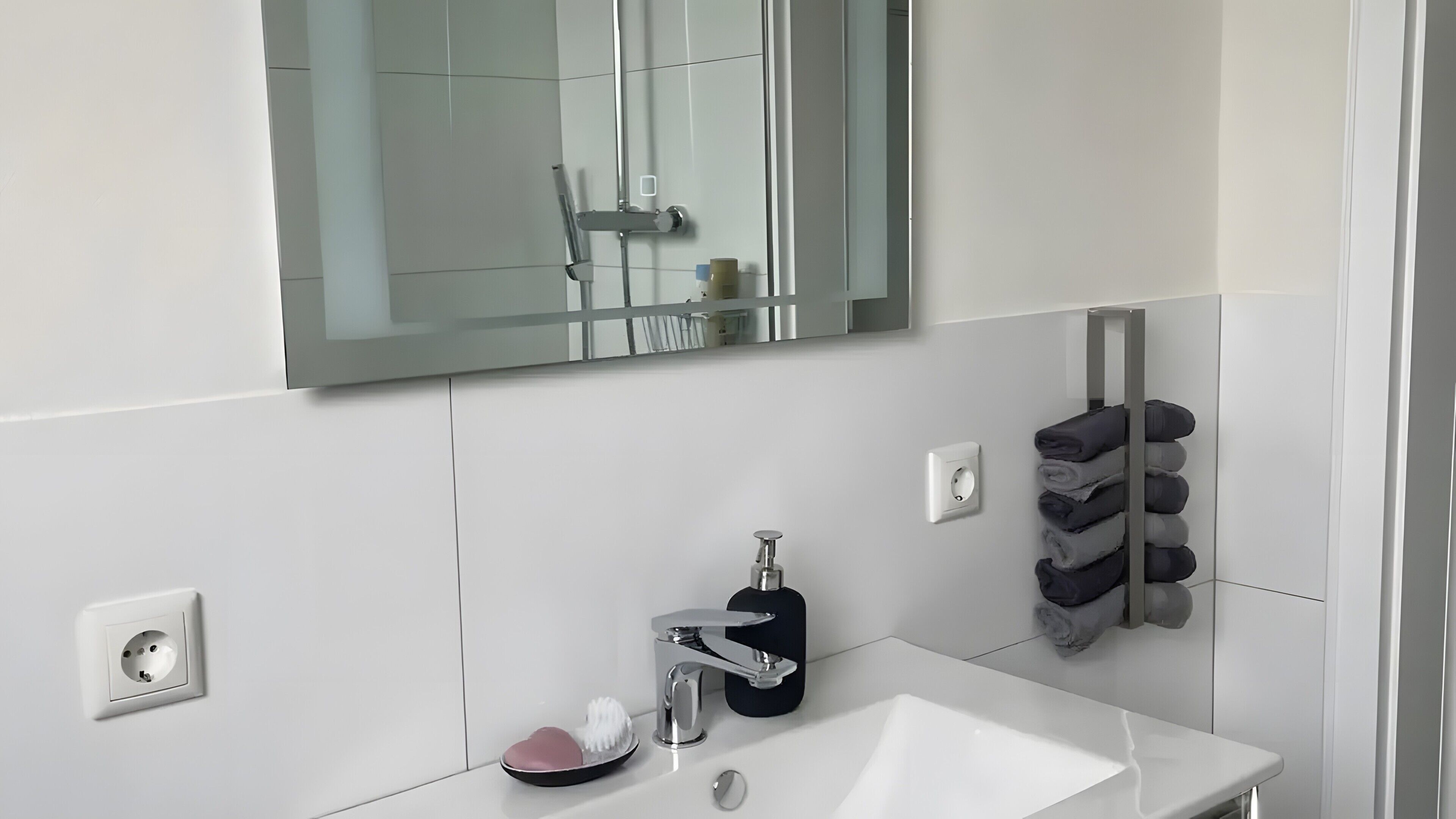 Un baño moderno con un lavamanos blanco, un espejo, un perchero para toallas y un dispensador de jabón.