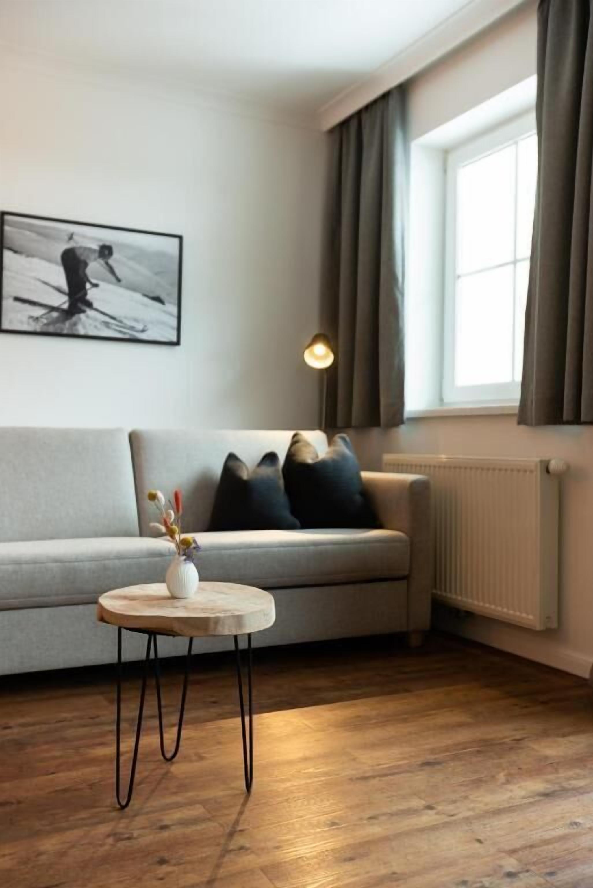 Una sala de estar con un sofá, una pequeña mesa redonda con un florero, una obra de arte blanco y negro en la pared y una ventana con cortinas.