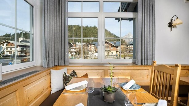 Ein Essbereich mit Tisch für zwei Personen, ein Fenster mit Blick auf Häuser und Bäume sowie eine hölzerne Bank mit Kissen.