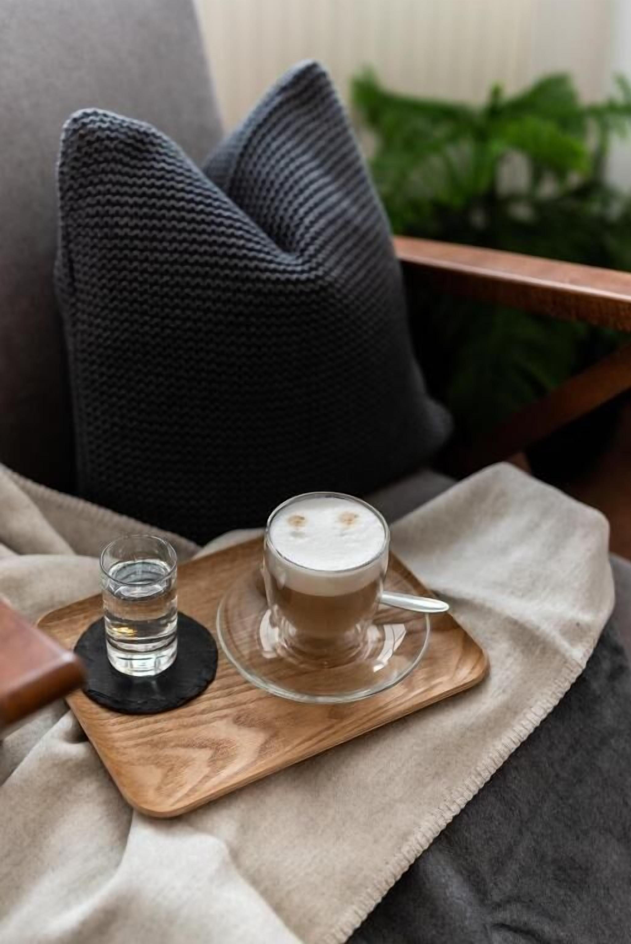 A wooden tray with a glass of coffee and a small glass of water, placed on a beige cloth.