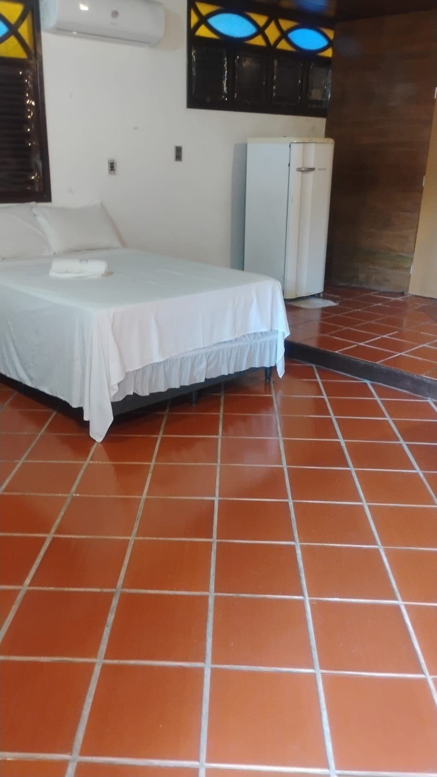 Una habitación con una cama, una pared blanca, un armario pequeño y un suelo de baldosas.