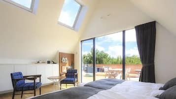 A bedroom with a large bed, two chairs, a small table, a wooden cabinet, and a view of the outdoors through large windows.
