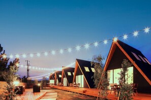 A row of triangular-roofed cabins with string lights overhead, wooden decks, and outdoor seating. - TENT Ichinomiya Glamping Resort (Ichinomiya)