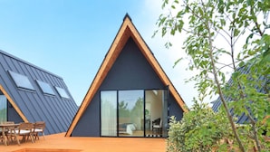 Una casa moderna con techo triangular, terraza de madera y puerta de vidrio que da al interior.