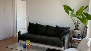 Una sala de estar con un sofá negro, una mesa de centro de vidrio, un sillón blanco, una planta grande en maceta y una mesita auxiliar con un libro y una cámara.