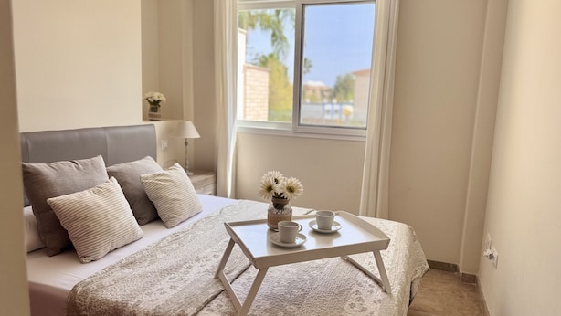 Quarto com cama, almofadas, uma mesinha com um vaso de flores e uma janela com vista.
