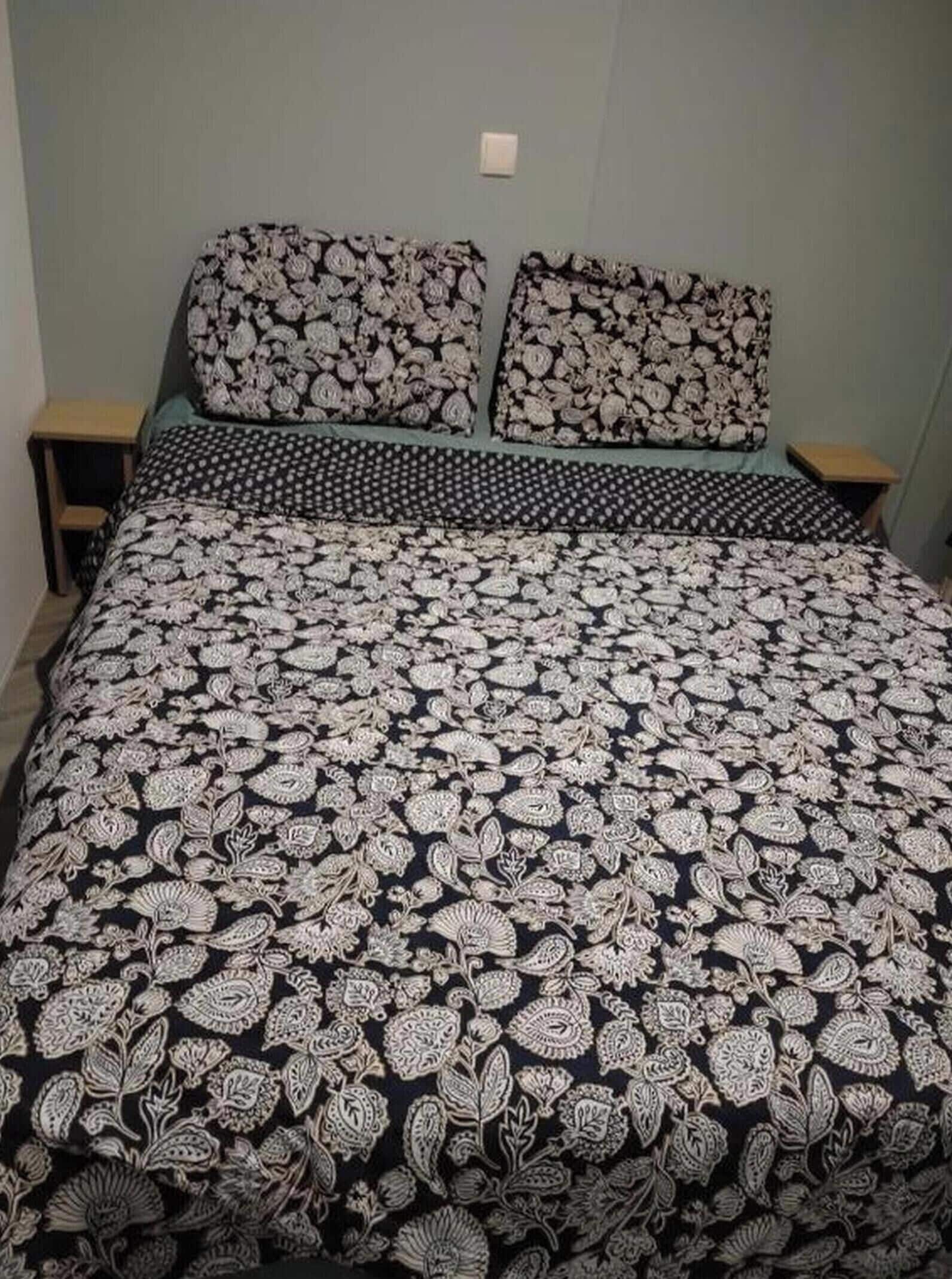 A bed with a patterned bedspread and matching pillows, two bedside tables, and a light-colored wall.