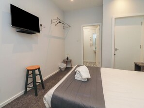 Quarto com uma cama, televisão na parede, um banquinho, mesa pequena com planta e porta que dá para um banheiro.