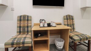 Ein Zimmer mit an der Wand montiertem Fernseher, zwei Karo-Stühlen, einem kleinen Holztisch, einem Mülleimer und einer Steckdose.