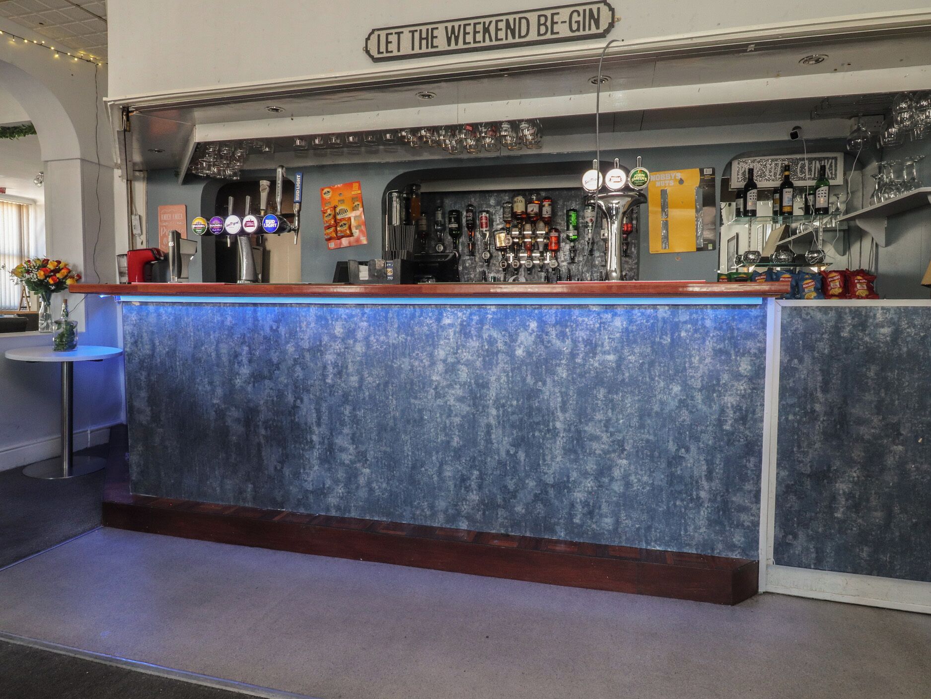 A bar area with a sign reading 'LET THE WEEKEND BE-GIN,' a selection of bottles, glasses, and a blue bar counter.