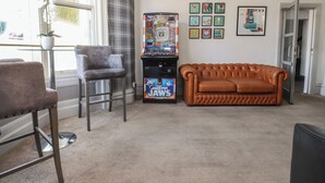 A room with a brown leather sofa, a grey armchair, a small table with a potted plant, and a slot machine. - Luxury Winter Gardens Apartment 2 (Blackpool)