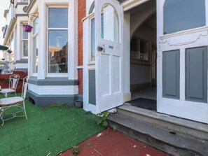 Un immeuble résidentiel doté d’une porte d’entrée, d’un petit espace extérieur aménagé avec des chaises, et d’une pelouse artificielle verte.