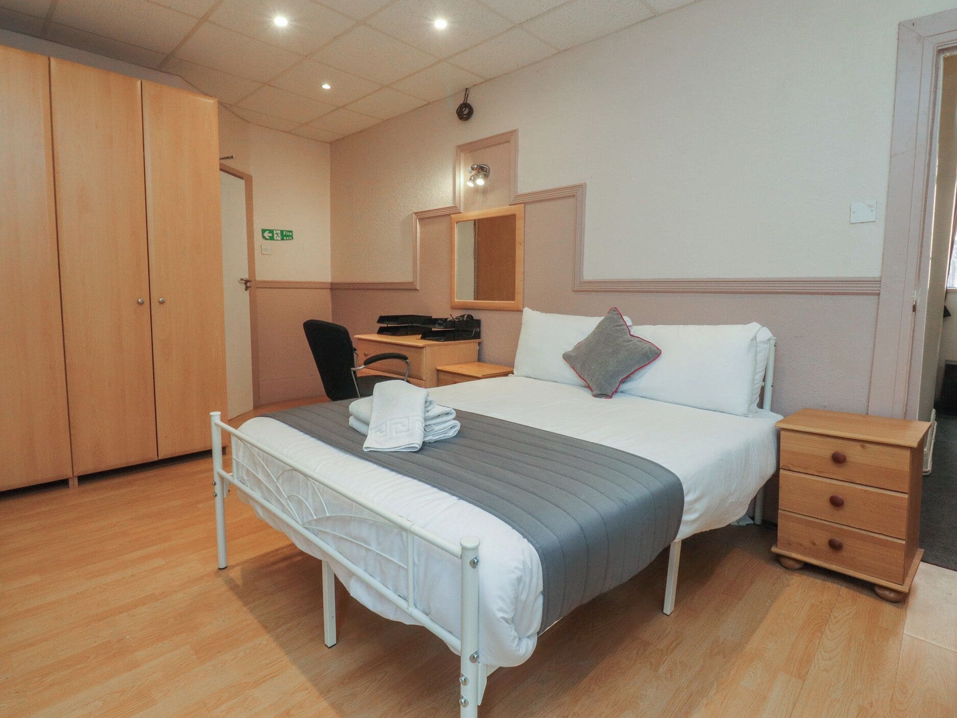 A hotel room with a bed, bedside table, wardrobe, desk, chair, and a mirror.