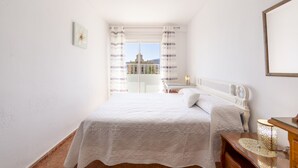 A bedroom with a bed, a bedside table, a lamp, and a view of a building through a window with curtains. - Almería Suite: Cathedral Penthouse (Almería)