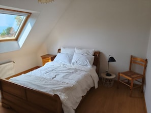 Makuuhuone, jossa on kalteva katto, puinen sänky, valkoiset vuodevaatteet, yöpöytä lampun kanssa ja tuoli.
