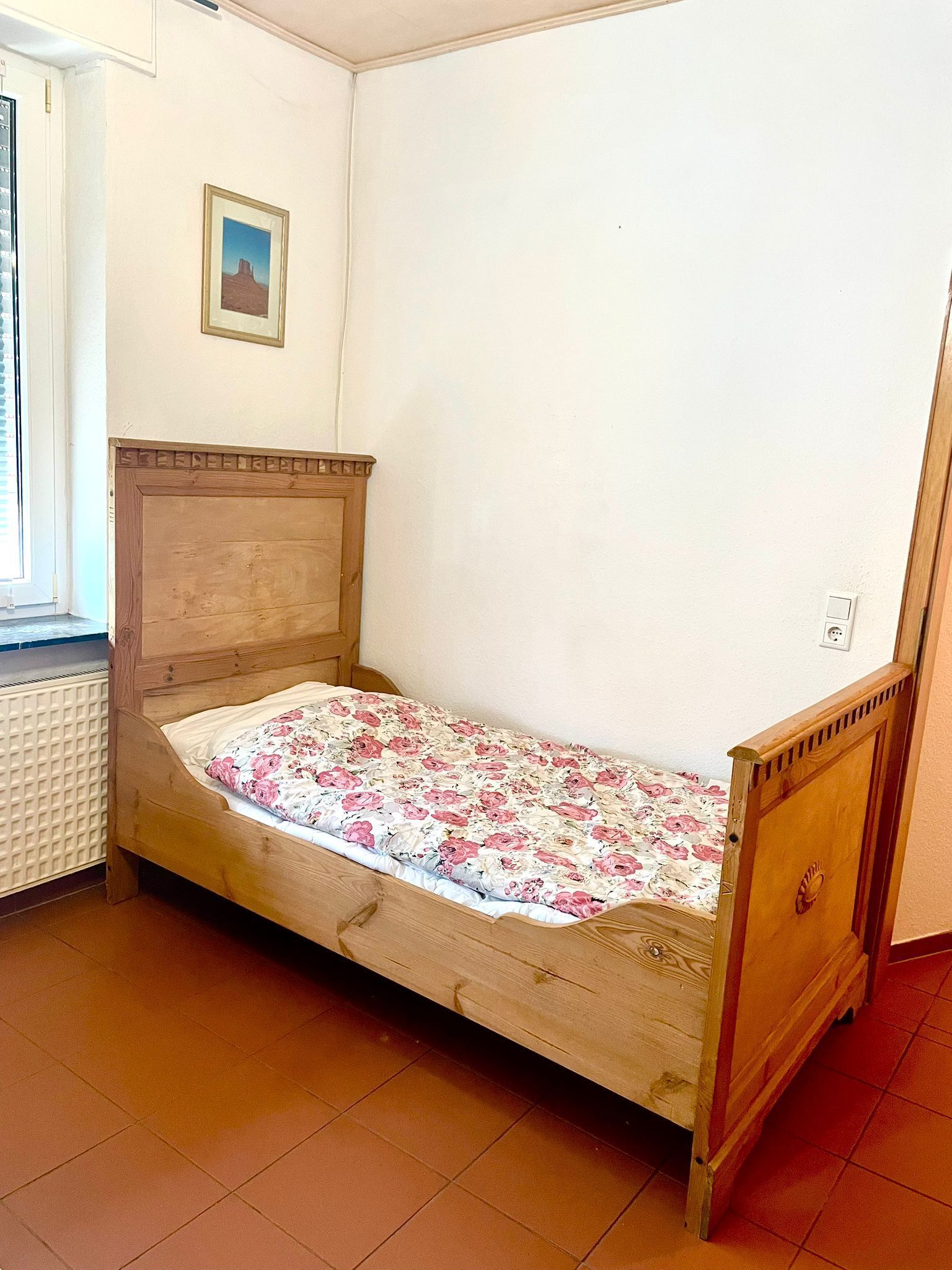 A single bed with a floral bedspread in a room with white walls, a radiator, and a small framed picture on the wall.
