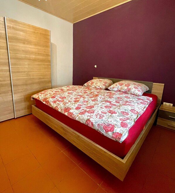 Un dormitorio con estructura de cama de madera, ropa de cama roja, una mesita de noche de madera y un armario.