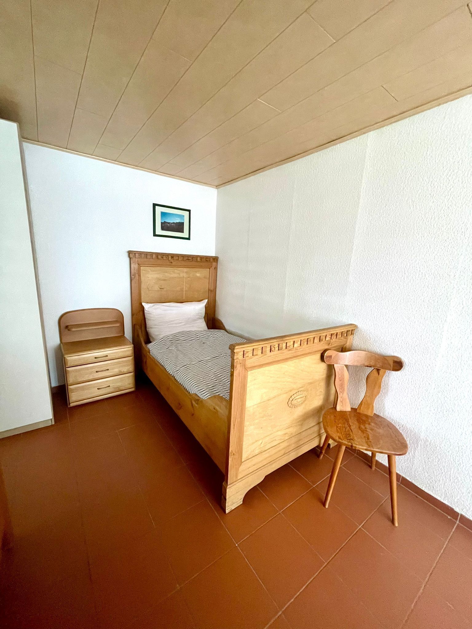 Een slaapkamer met een houten bed, een nachtkastje van hout, een stoel en een schilderij aan de muur.