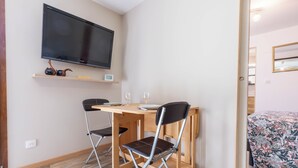 Ett litet rum med en platt-TV som är monterad på väggen, ett träbord med två stolar och en säng med ett blommigt överkast.
