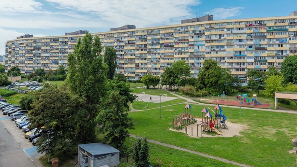 Ein mehrstöckiges Wohngebäude mit Balkonen, ein Spielplatz mit Rutschen und Schaukeln sowie ein Parkplatz mit Autos.