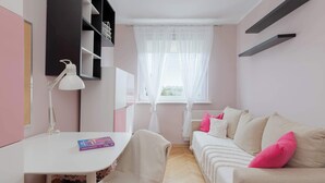 Ein Zimmer mit einem weißen Schreibtisch, einem weißen Stuhl, einem beigen Sofa mit rosafarbenen Kissen, einer weißen Lampe und einem Fenster mit durchsichtigen Vorhängen.