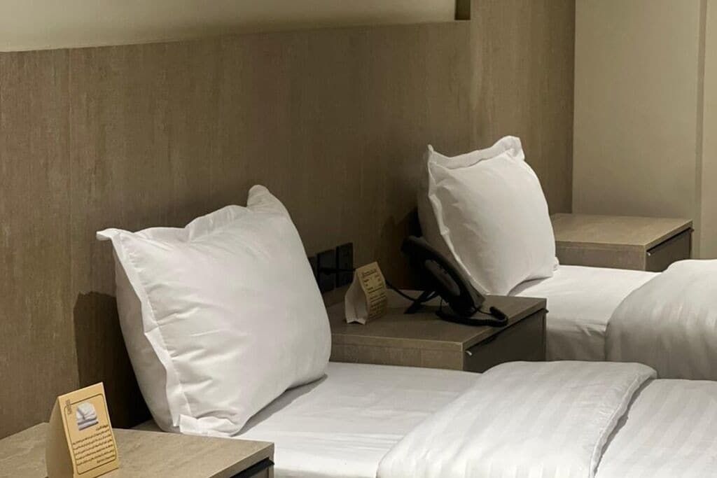 A hotel room with two beds, each with white bedding and pillows. There is a bedside table with a lamp and a small card. The room has a wooden headboard and a neutral color scheme.