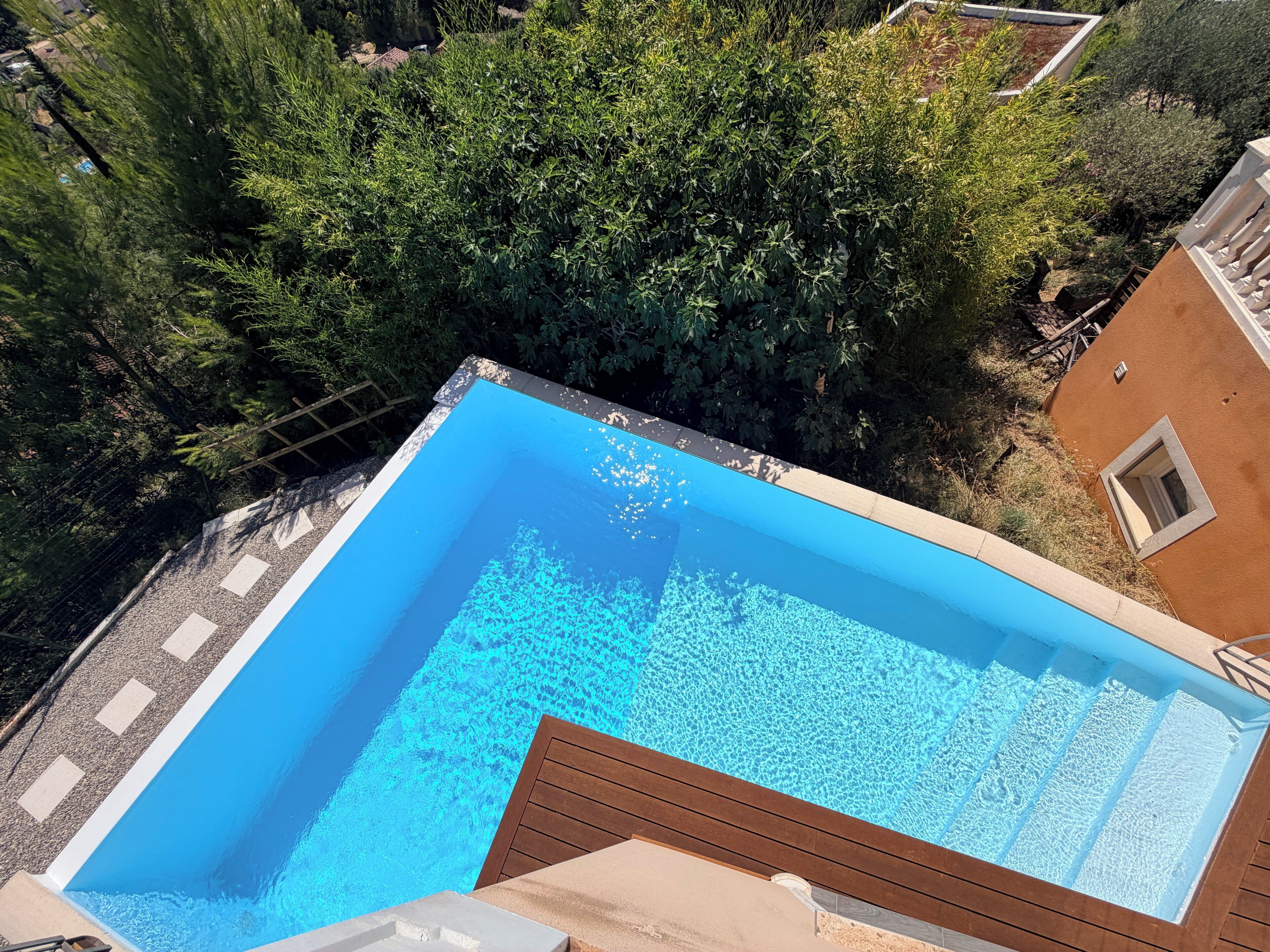 Una piscina rectangular con agua azul cristalina, rodeada por una terraza de madera y un jardín con diversas plantas.