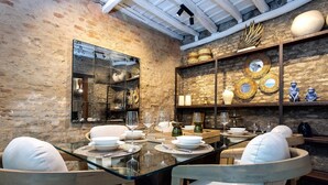 Uma área de jantar com mesa de vidro, cadeiras acolchoadas brancas e parede de tijolos ao fundo.