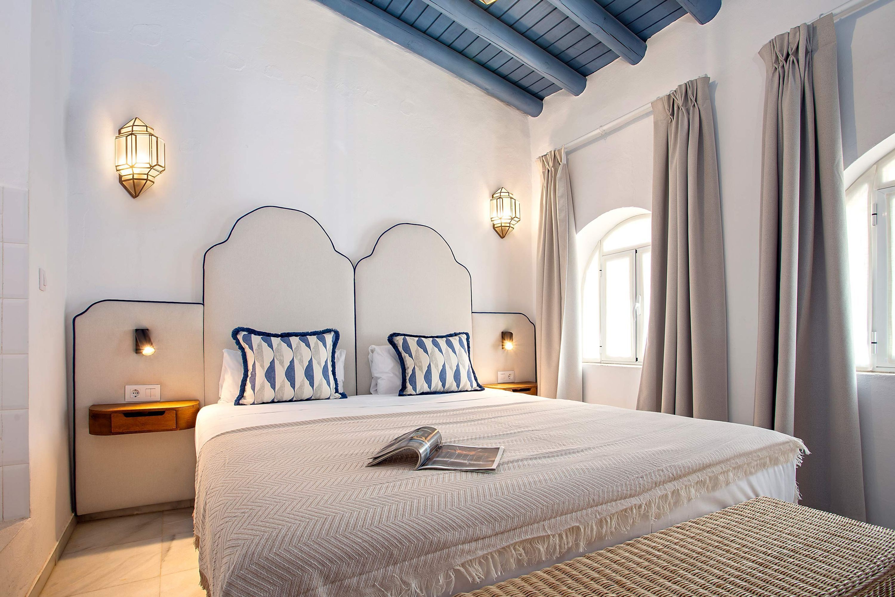 A neatly made bed with a patterned bedspread, two bedside tables with wall-mounted lamps, and a window with curtains.