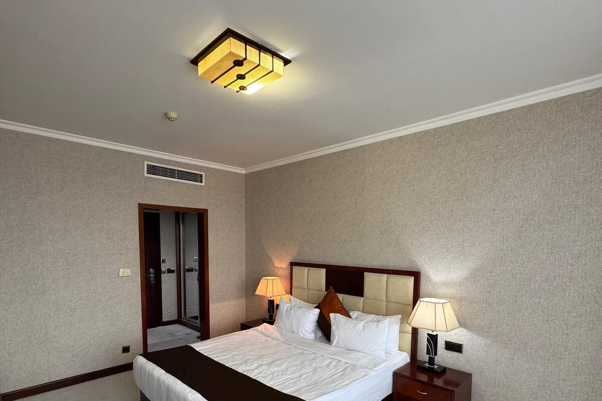 Una habitación de hotel con una cama grande, lámparas de noche, un cabecero, una mesita de noche y una puerta.