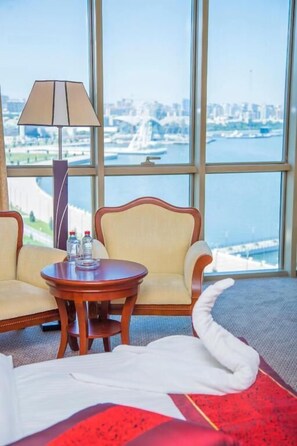 Habitación de hotel con cama, dos sillas, una mesa con lámpara y vistas a un paisaje urbano a través de amplias ventanas.