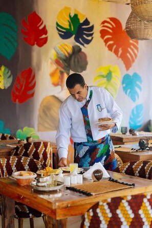 A person serving food at a wooden table with a colorful tropical leaf mural in the background. - Ô TALIA Hôtel and SPA (Nungwi)
