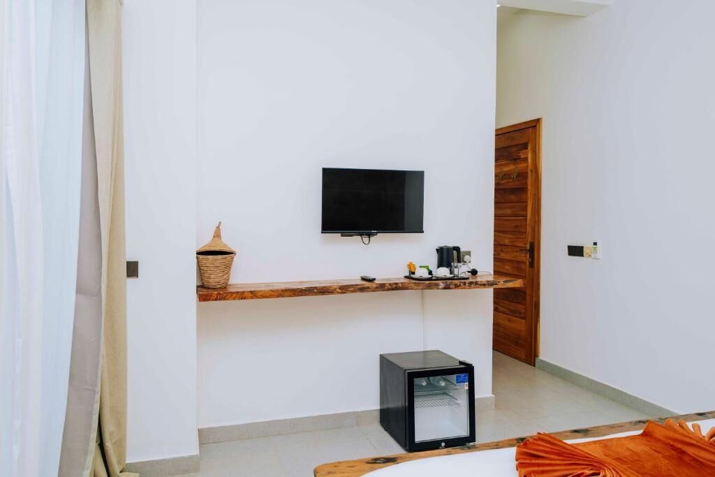 A room with a flat-screen TV mounted on the wall, a small refrigerator, a wooden shelf with a basket, and a wooden door.