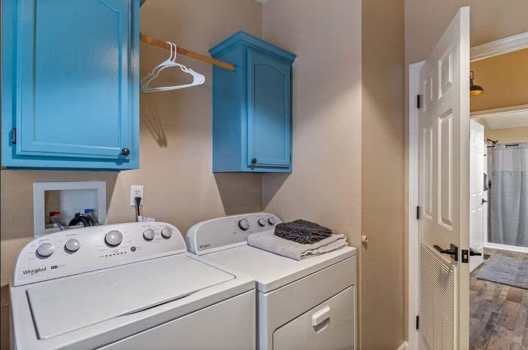 Une buanderie équipée de deux lave-linge et deux sèche-linge Whirlpool blancs, d’un meuble bleu et d’une porte blanche donnant accès à une salle de bain.