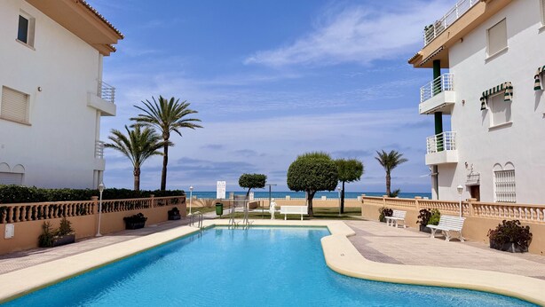 Una piscina amb vista despejada al mar, envoltada de palmeres i un edifici blanc amb balcons.